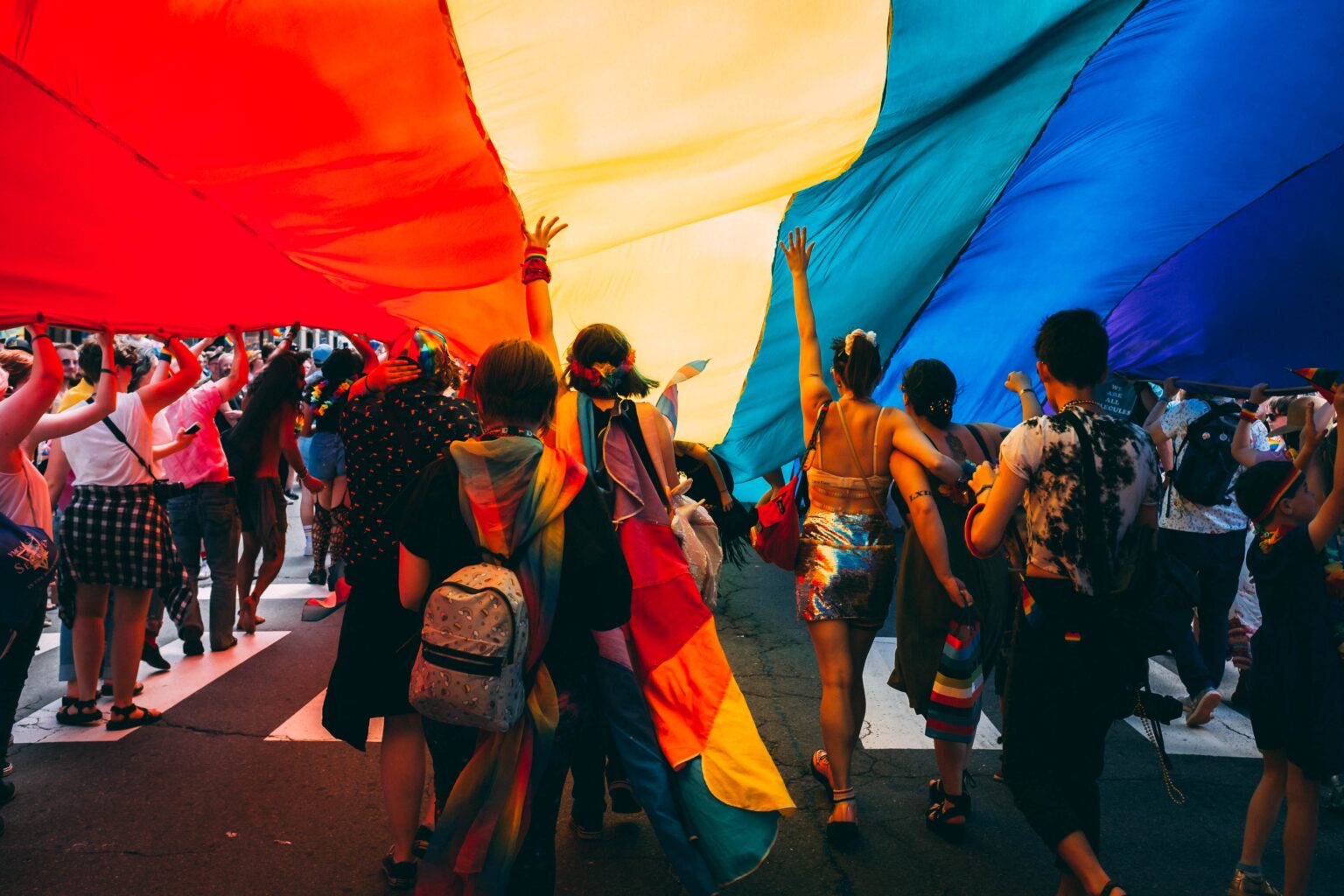 A group of people seen from the back marching under a rainbow flag