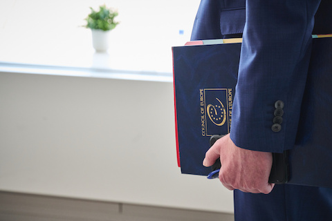 A person in a blue suit carries a notebook with the Council of Europe logo. We only see the notebook, hand and arm, partially.