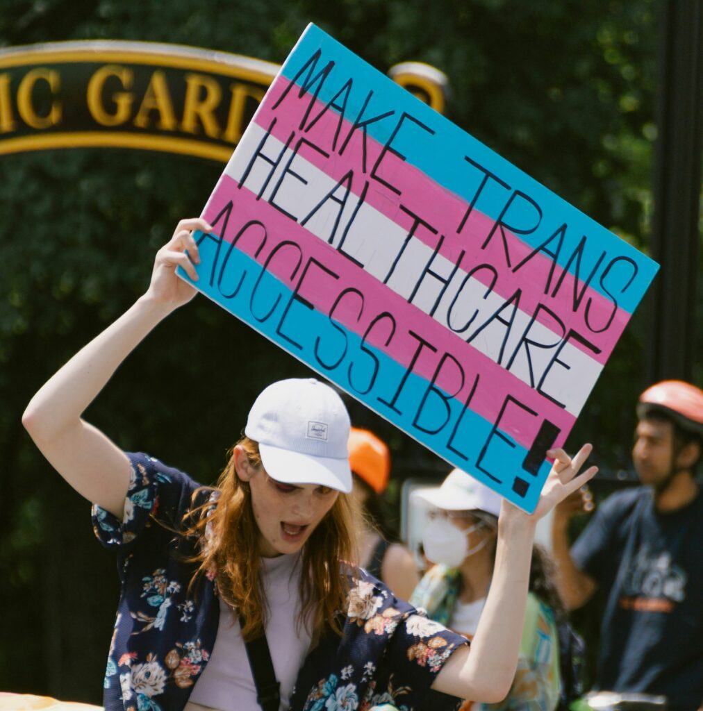 A person holding a sign with trans flag colours as background, saying "MAKE TRANS HEALTHCARE ACCESSIBLE"