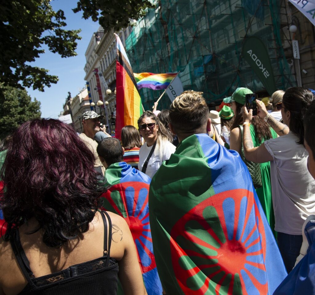 People at a Pride March holding and wearing rainbow and Roma flags.