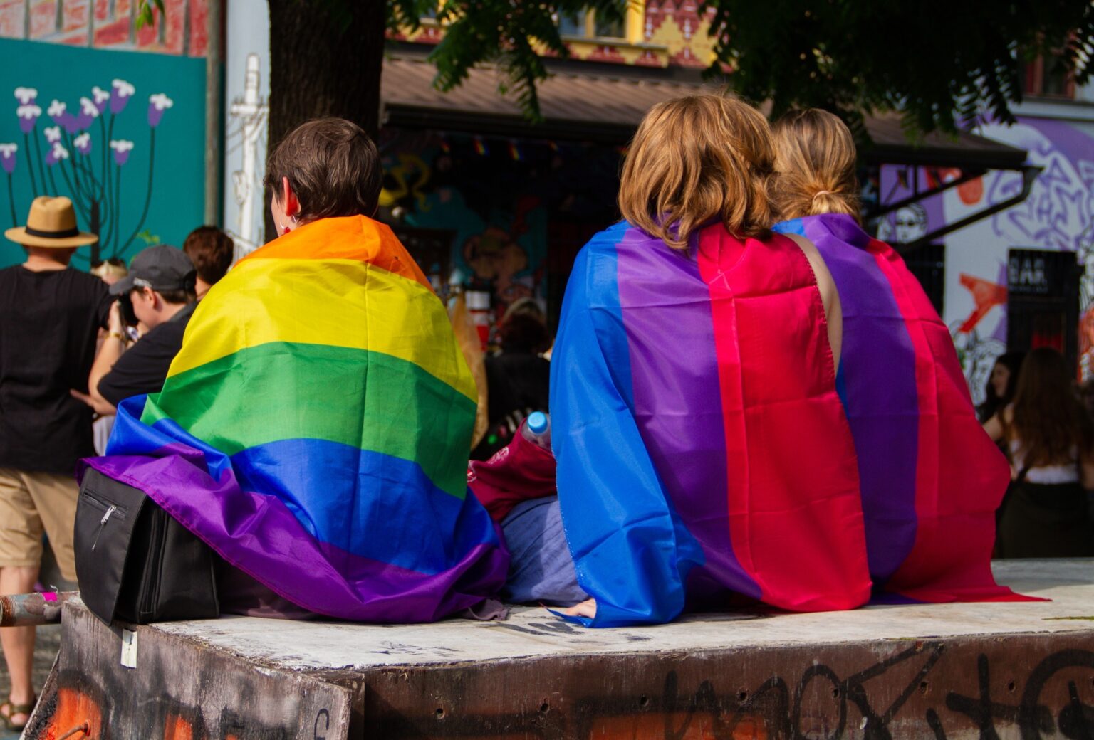 Three people sitting together, one person is wrapped in a rainbow flag, while the other two are wrapped in bi+ pride flags,