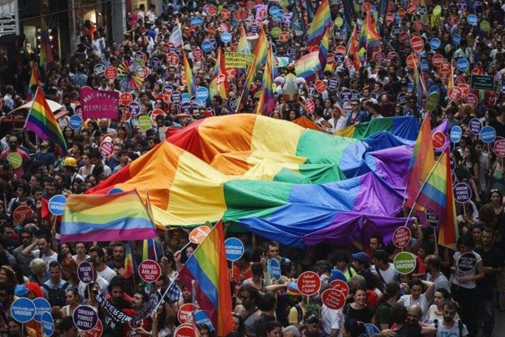 A photo of a large gathering with a massive rainbow flag is stretched over the participants